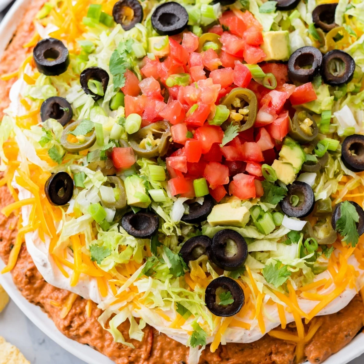 A close-up of Ultimate Layered Taco Dip shows melted cheddar, black olives, and sliced jalapeños beside a basket of crunchy tortilla chips.