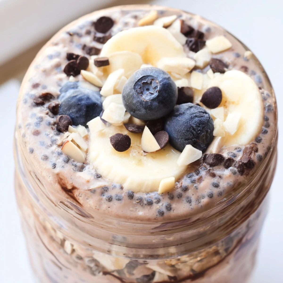 A close-up of Ultimate Healthy Chocolate Overnight Oats in a clear jar, topped with banana slices and chopped almonds.