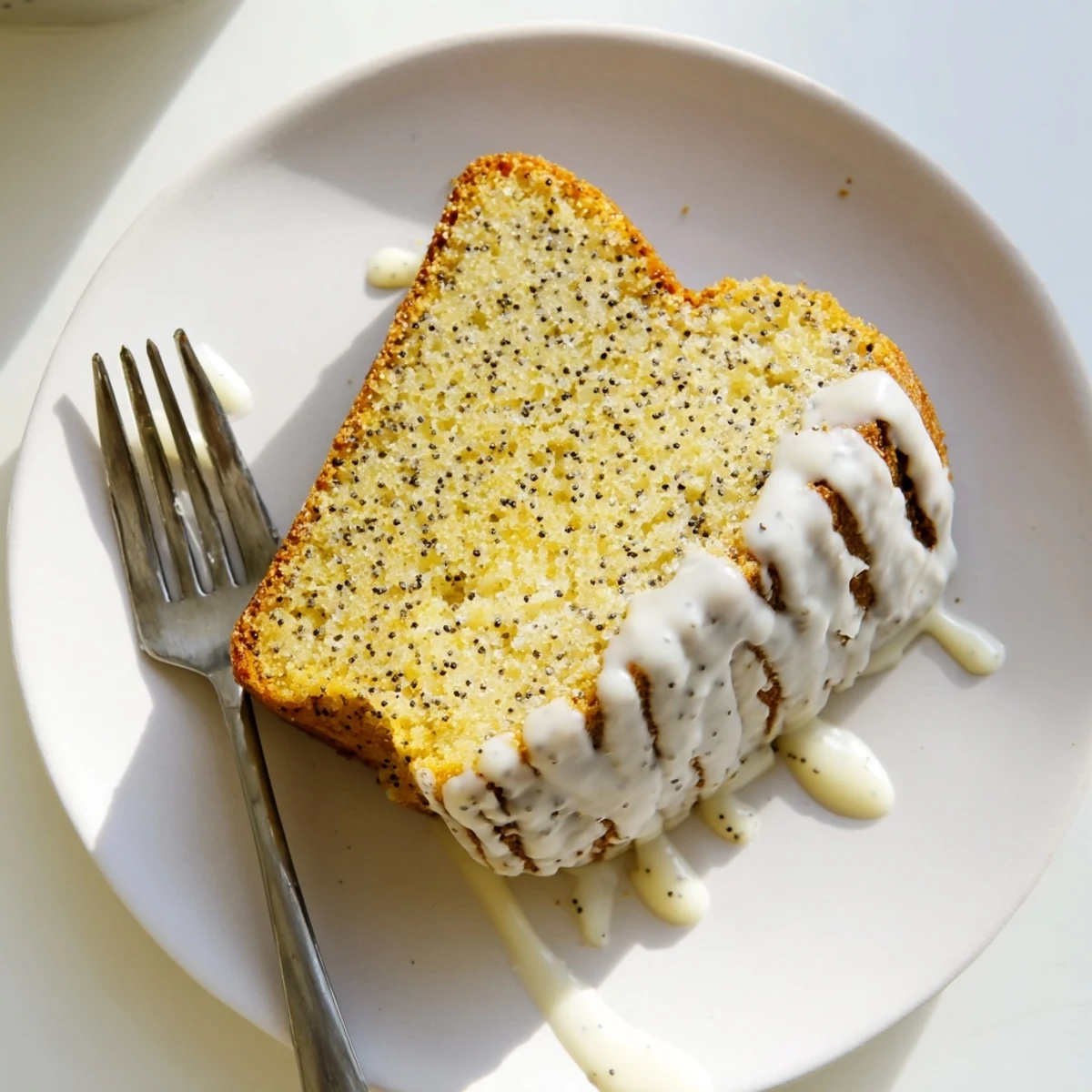 A slice of glazed Lemon Poppy Seed Cake rests on a white plate, showcasing its moist crumb and poppy speckles.