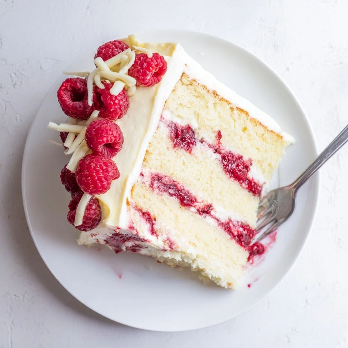 Elegant slice of white chocolate raspberry dream cake topped with fresh raspberries and chocolate curls